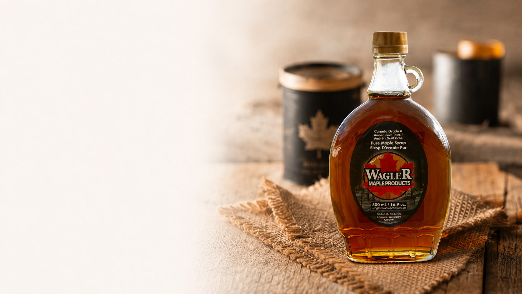 A bottle of Wagler Maple Products maple syrup stands on a rustic burlap cloth on a wooden table, with two dark metal canisters blurred in the background.