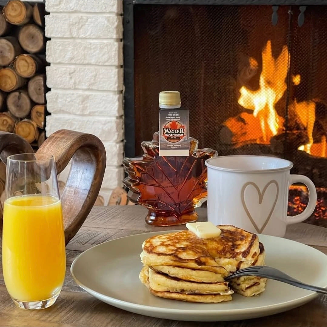Stack of pancakes served with Wagler Maple Product's pure Ontario maple syrup with a glass of orange juice and a mug in front of a fireplace