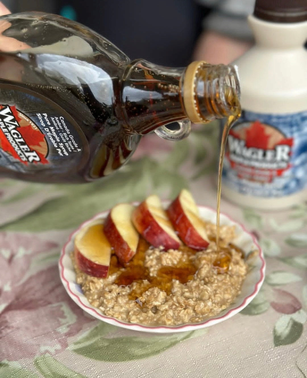 Wagler's pure Ontario maple syrup being poured over a bowl of oatmeal with apple slices on a floral tablecloth.