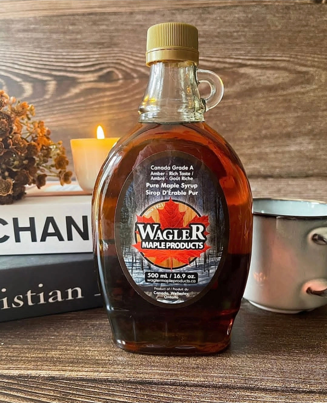 Glass bottle of Wagler Maple Products pure Ontario maple syrup on a wooden surface with a candle and books in the background.